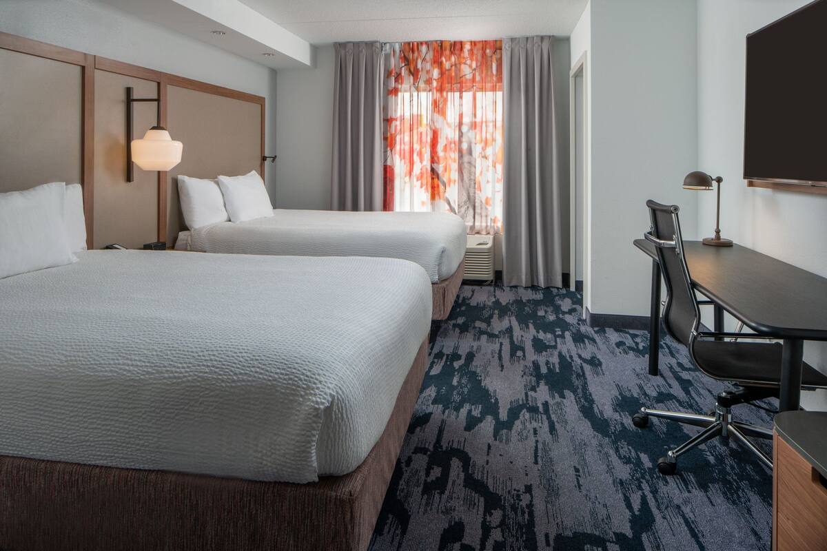 A hotel room with two beds, a desk, a chair, and a large window with curtains.