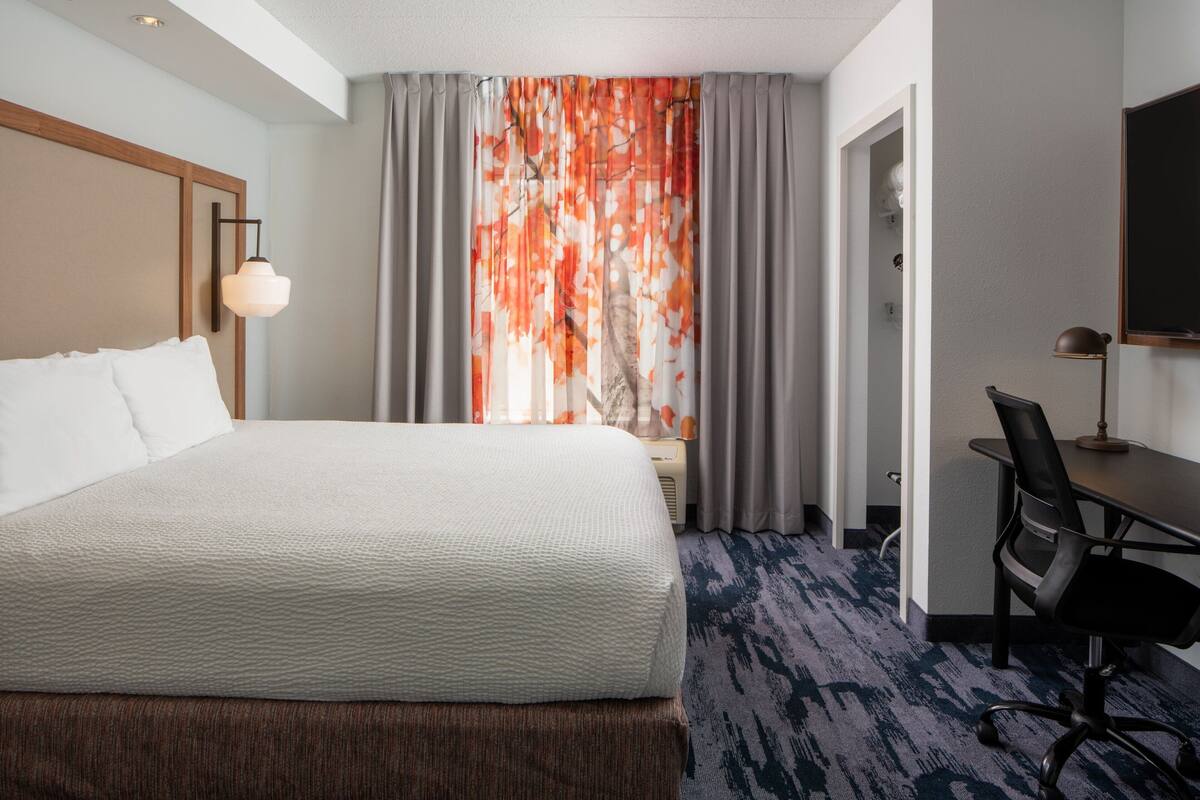 A hotel room with a large bed, a desk, and a chair. There is a window with curtains, a lamp, and a television on the wall.