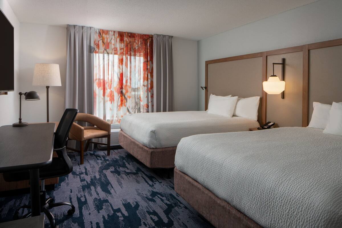 A hotel room with two beds, a desk, a chair, a lamp, and a window with curtains.
