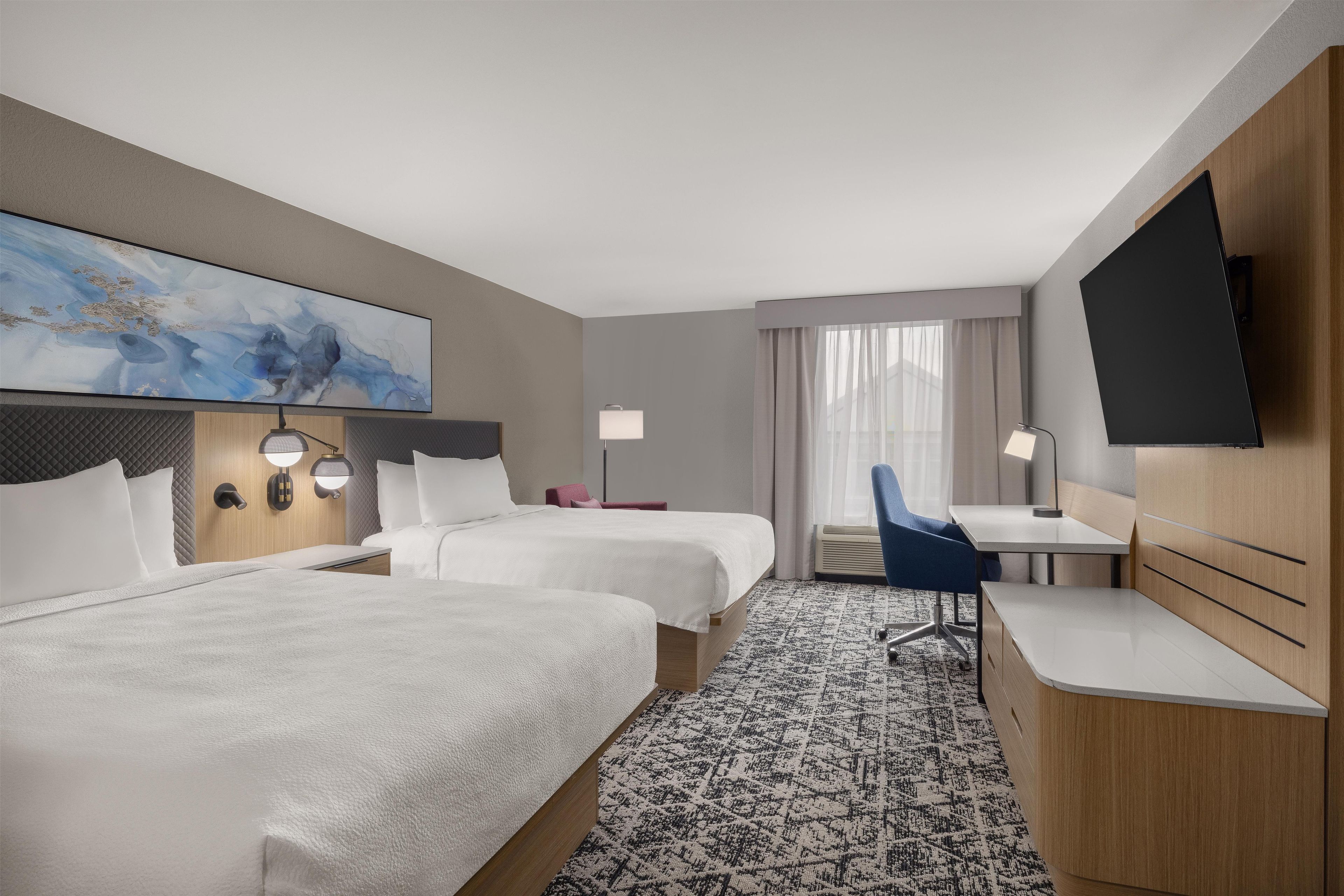 A hotel room with two beds, a desk, a chair, a television, and a painting on the wall.