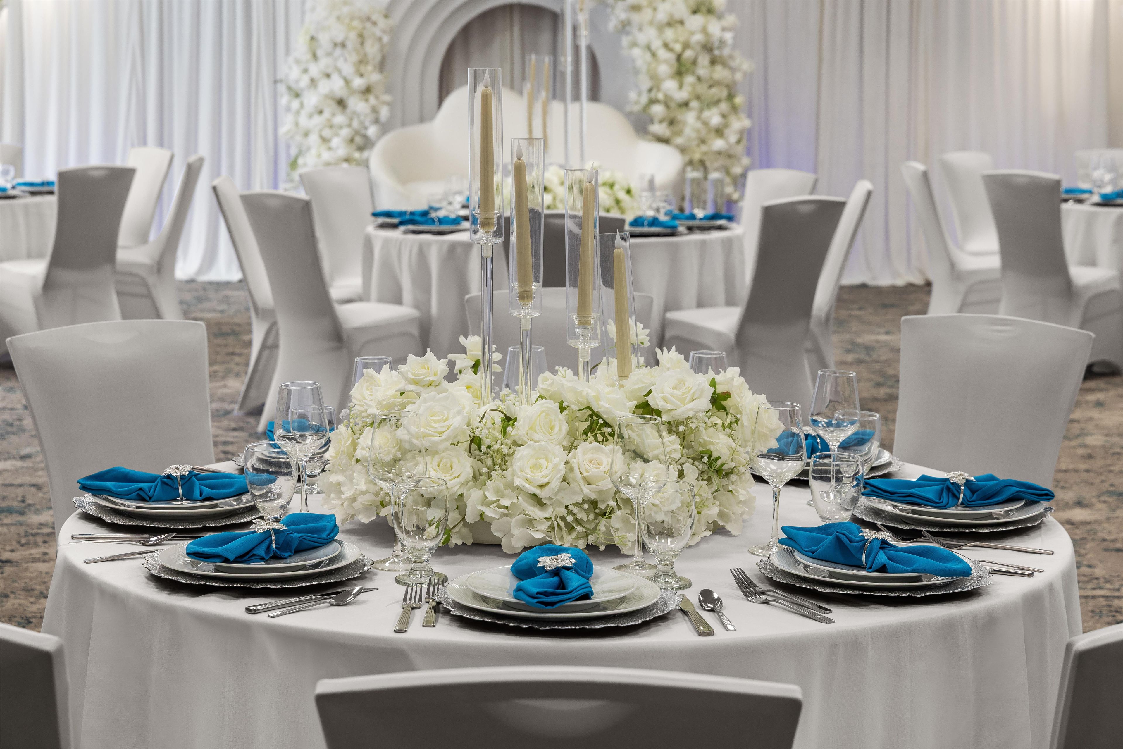 Mesa redonda dispuesta para un evento formal, con arreglos florales blancos, cristalería transparente y servilletas azules.