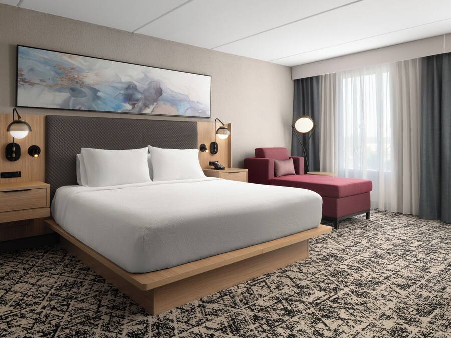 Una habitación de hotel moderna con una cama grande, un sillón rojo, un cuadro en la pared y vistas al exterior a través de la ventana.