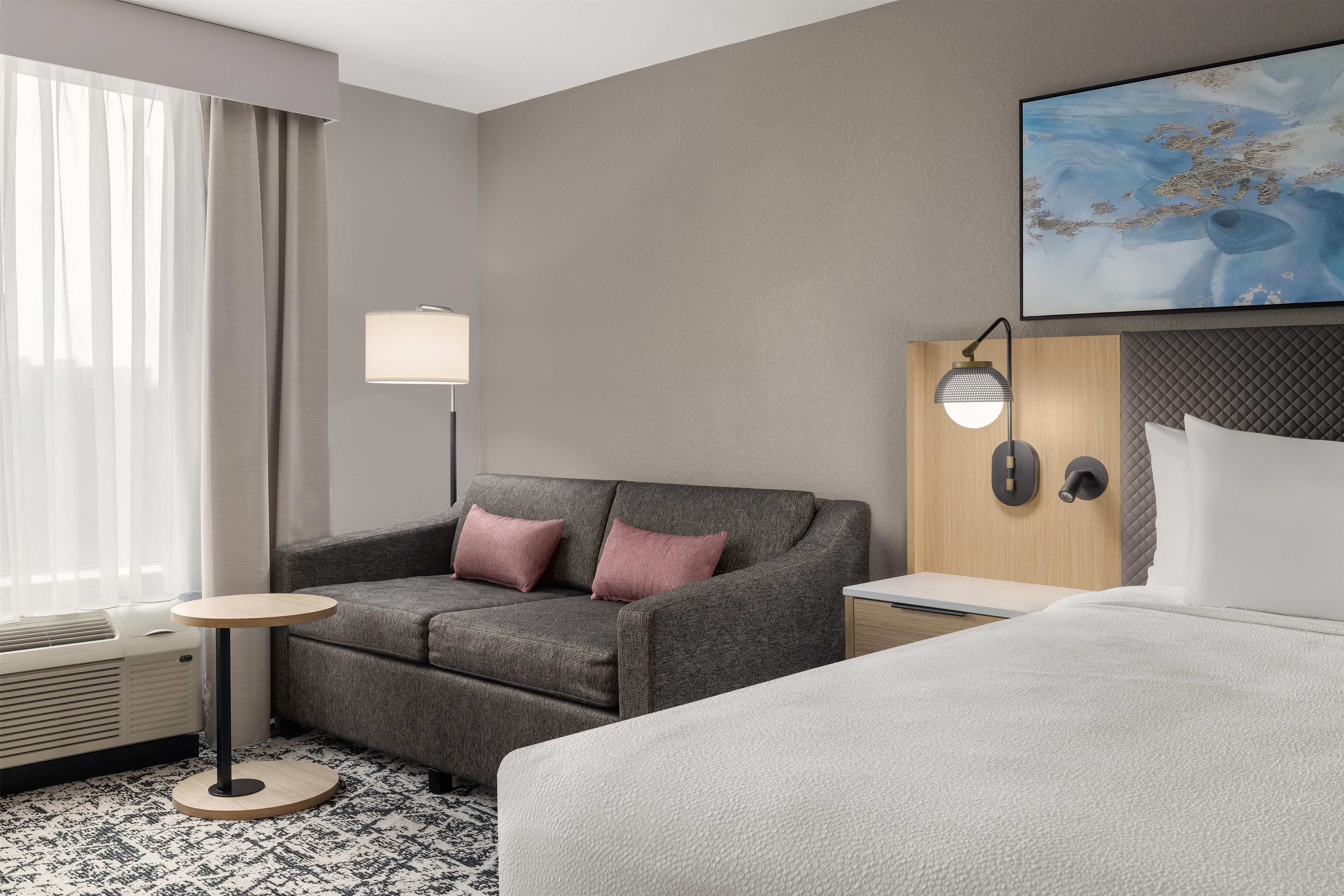 A hotel room with a grey sofa, a small round table, a bed, a standing lamp, and a painting on the wall.