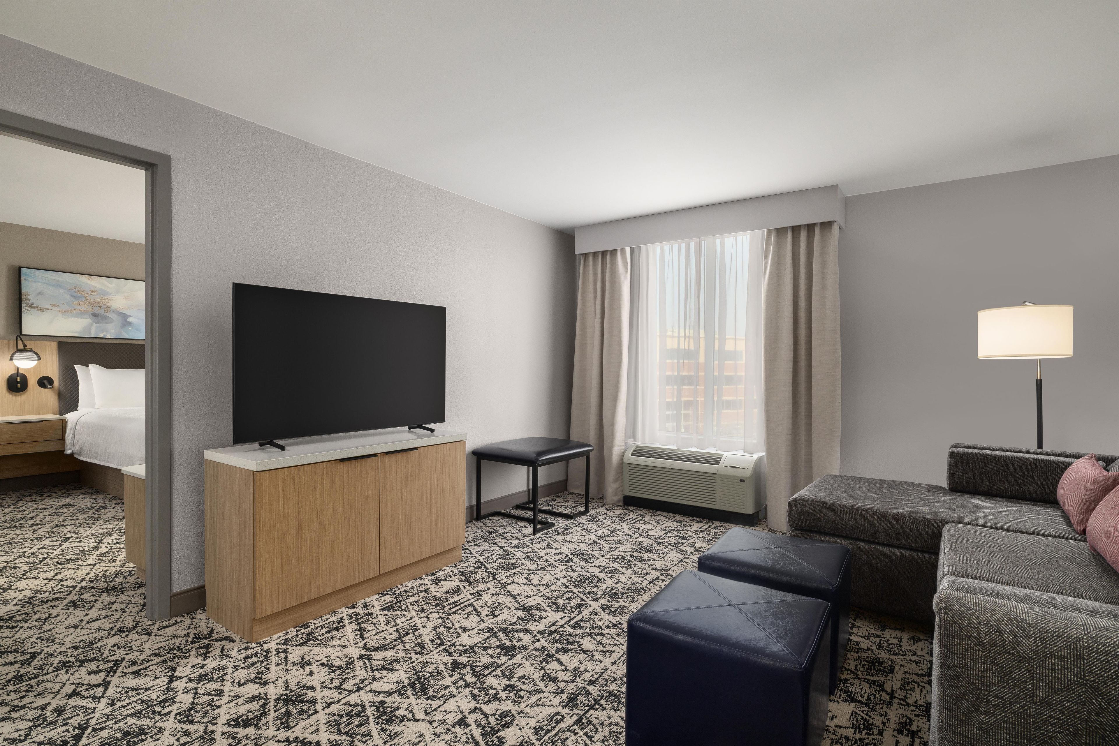 Ein Hotelzimmer mit Bett, Fernseher, Schreibtisch, Stuhl, Sofa und einem Fenster mit Vorhängen.