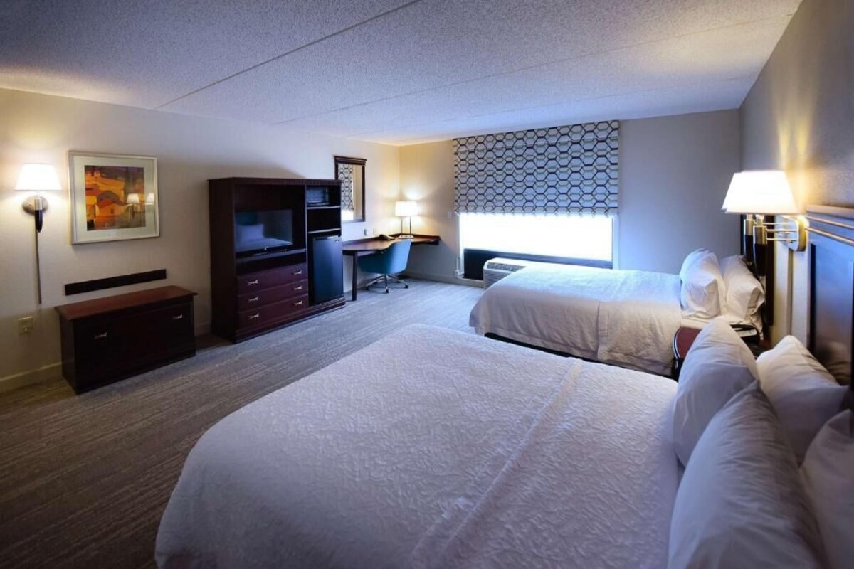 A hotel room with two beds, a desk, a chair, a dresser, and a TV.