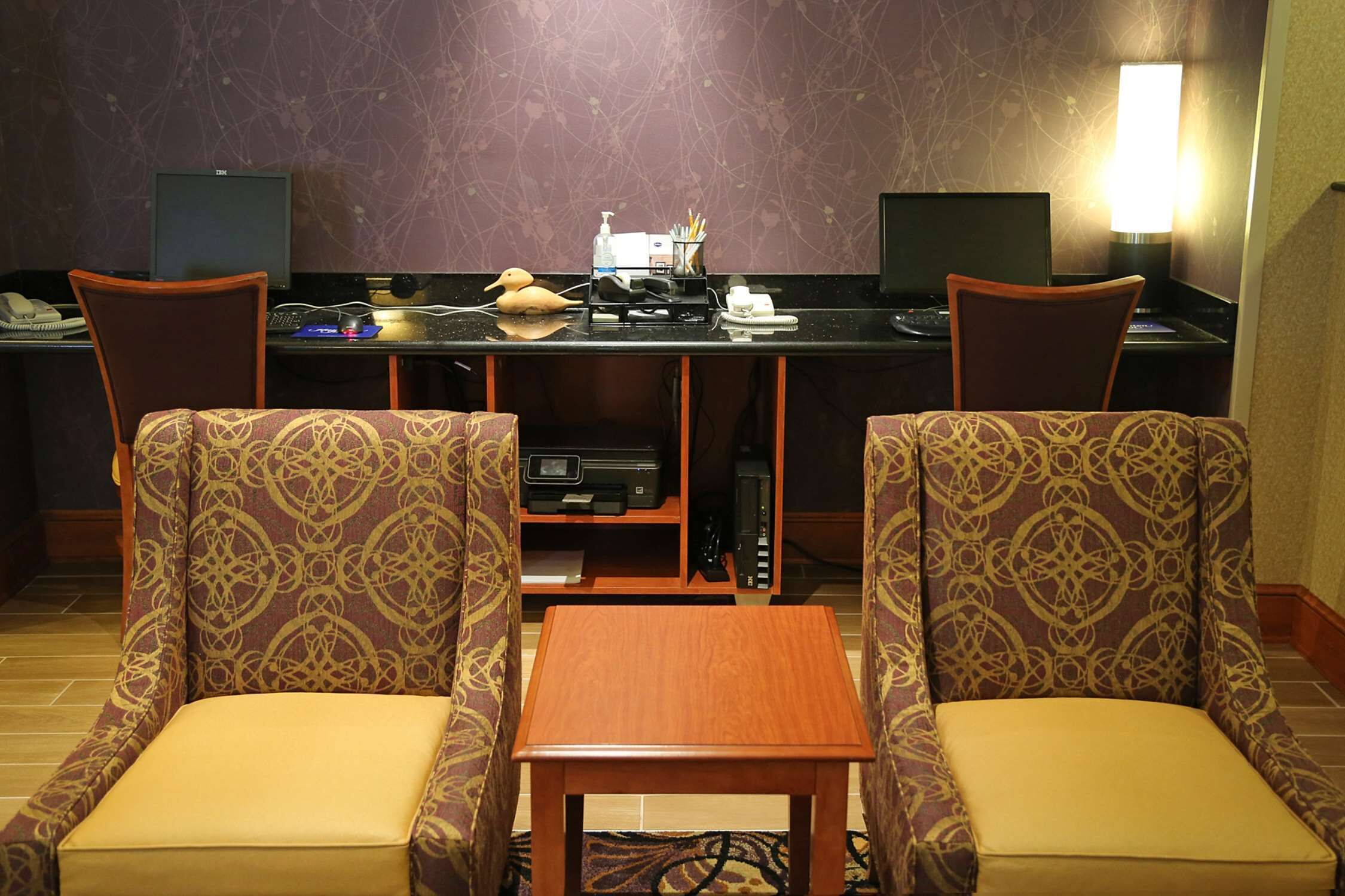 A hotel room with a desk, two patterned chairs, a small wooden table, and a lamp.