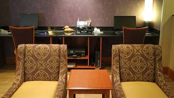 A hotel room with a desk, two patterned chairs, a small wooden table, and a lamp.