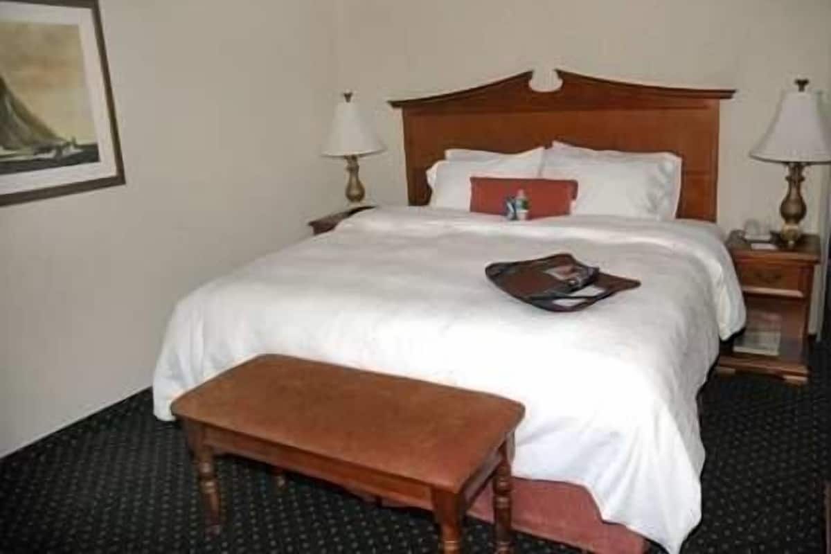 Quarto de hotel com cama, mesa de cabeceira, luminária, banquinho e um quadro na parede.