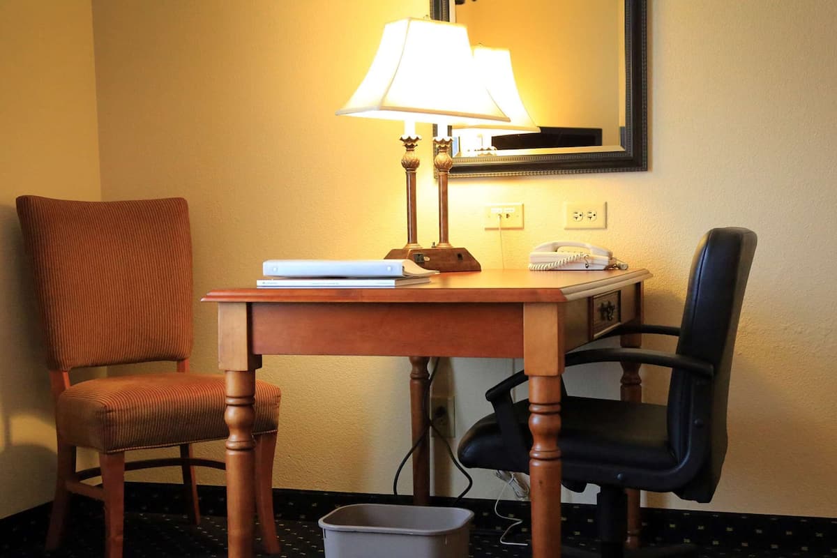 Quarto de hotel com escrivaninha de madeira, duas cadeiras, luminária, telefone e lixeira.