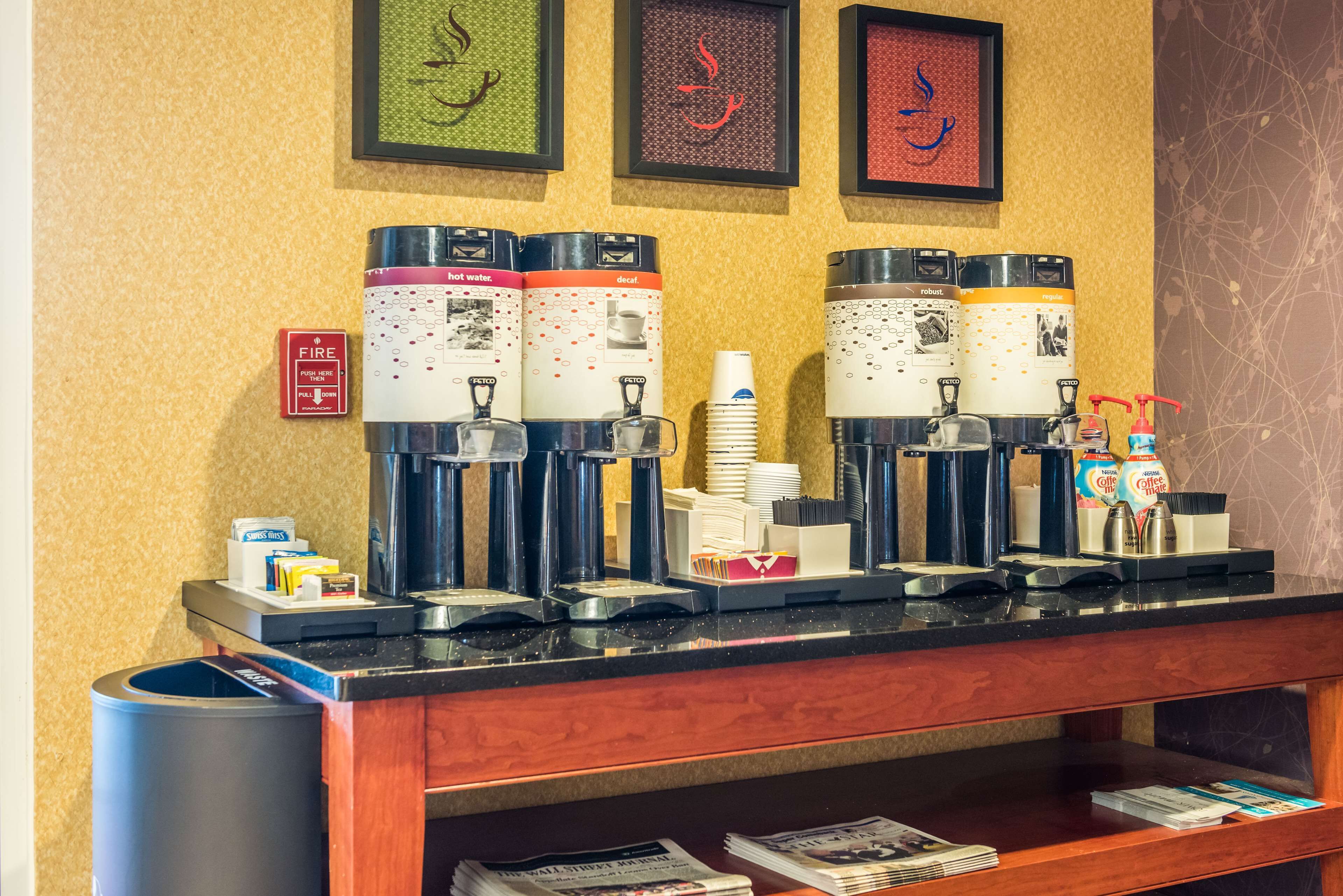 A coffee station with multiple dispensers, a fire alarm, and framed artwork on the wall.