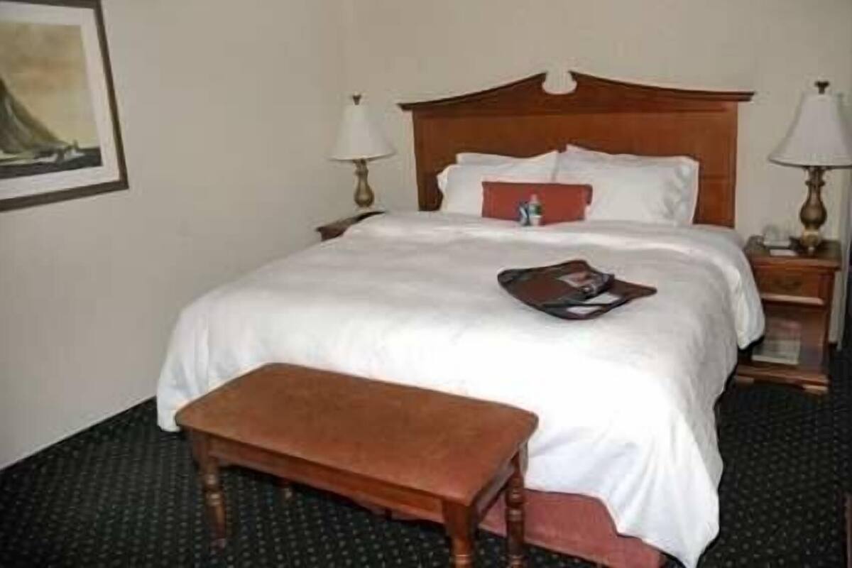 Quarto de hotel com cama, mesa de cabeceira, luminária, banquinho e um quadro na parede.