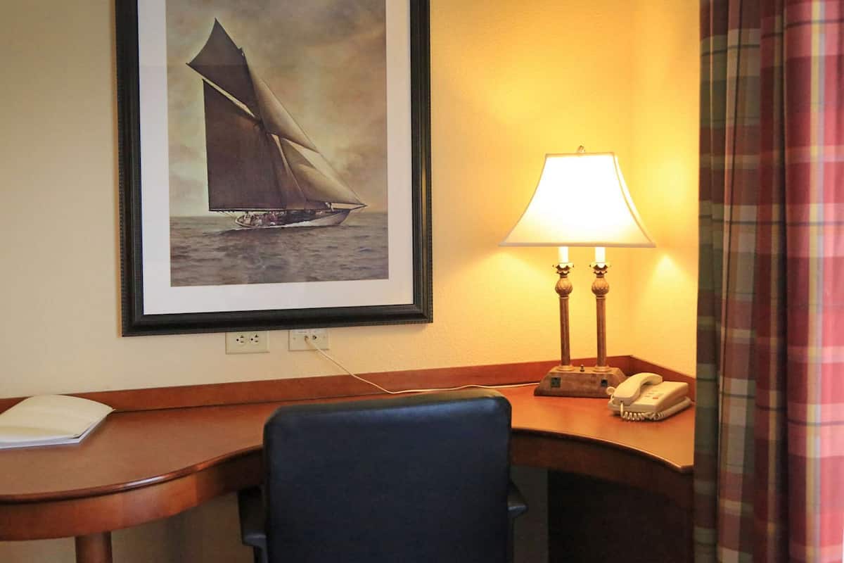 Quarto de hotel com uma escrivaninha, um quadro com um veleiro, uma luminária e um telefone.