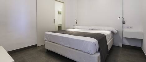 A modern bedroom with a bed, a bedside table, a mirror, and a wall-mounted light fixture.
