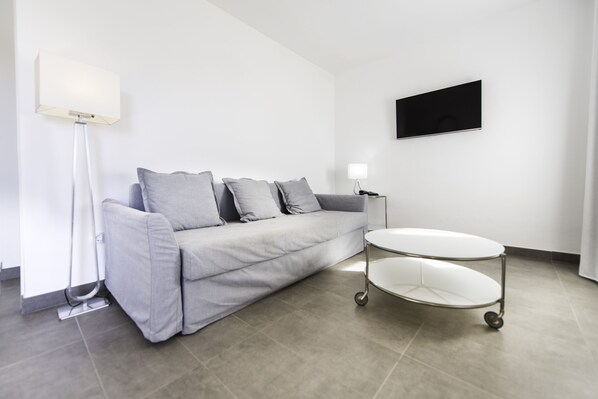 Una sala moderna con un sofá gris, una lámpara de pie blanca, un televisor de pantalla plana montado en la pared y una pequeña mesa blanca con ruedas.