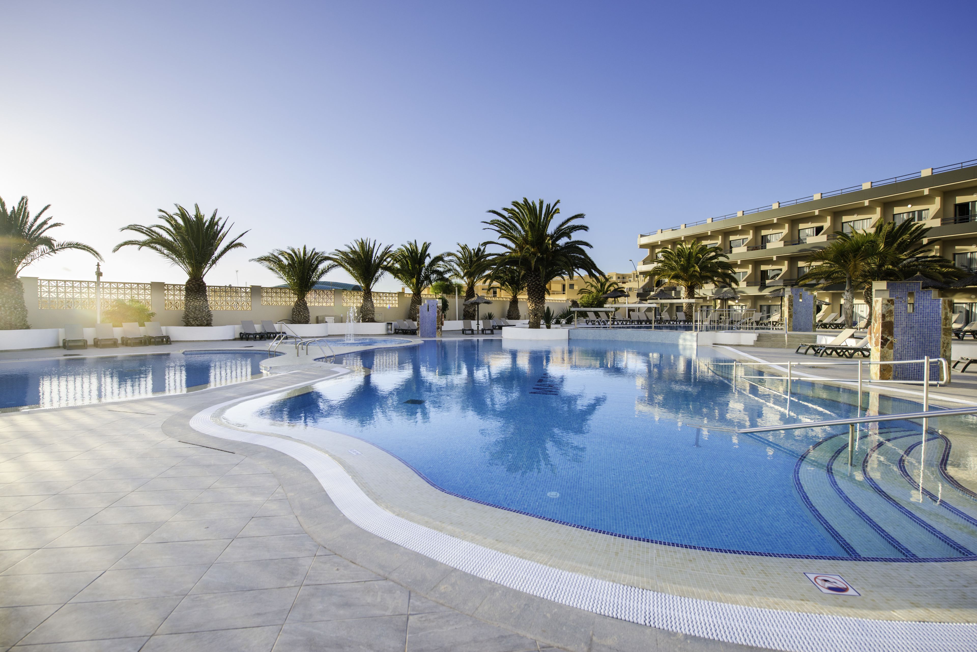 A swimming pool with a tiled deck, palm trees, and a building with balconies in the background.