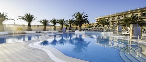 A swimming pool with a tiled deck, palm trees, and a building with balconies in the background.