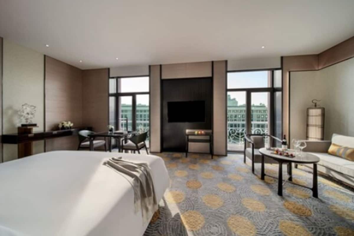 A modern hotel room with a large bed, a TV, a seating area with a table and chairs, and large windows offering a city view.