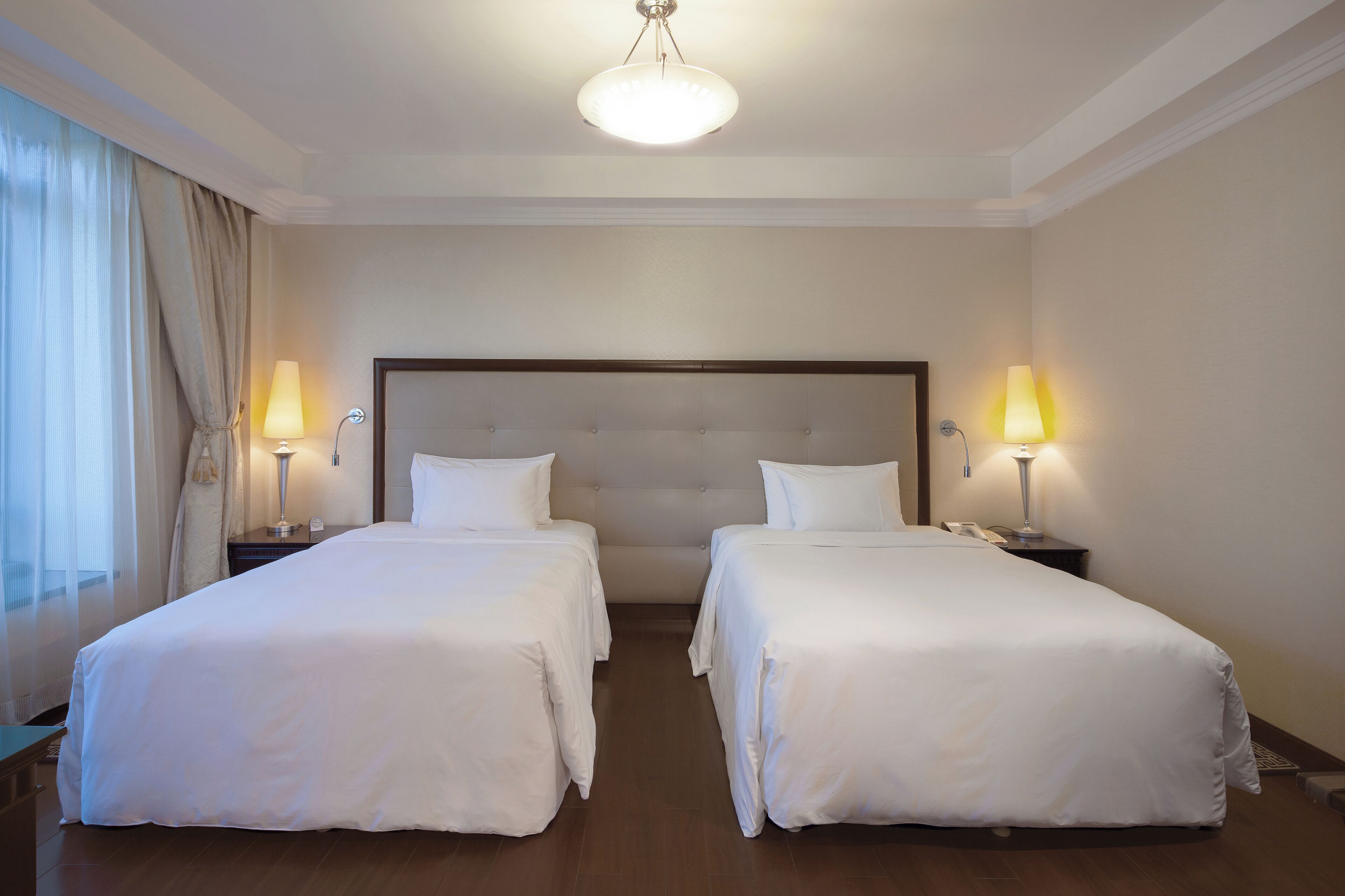 A hotel room with two single beds, a large padded headboard, bedside lamps, and a large window with curtains.