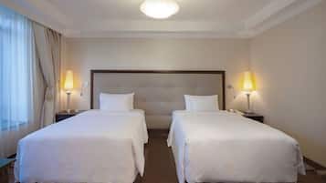 A hotel room with two single beds, a large padded headboard, bedside lamps, and a large window with curtains.