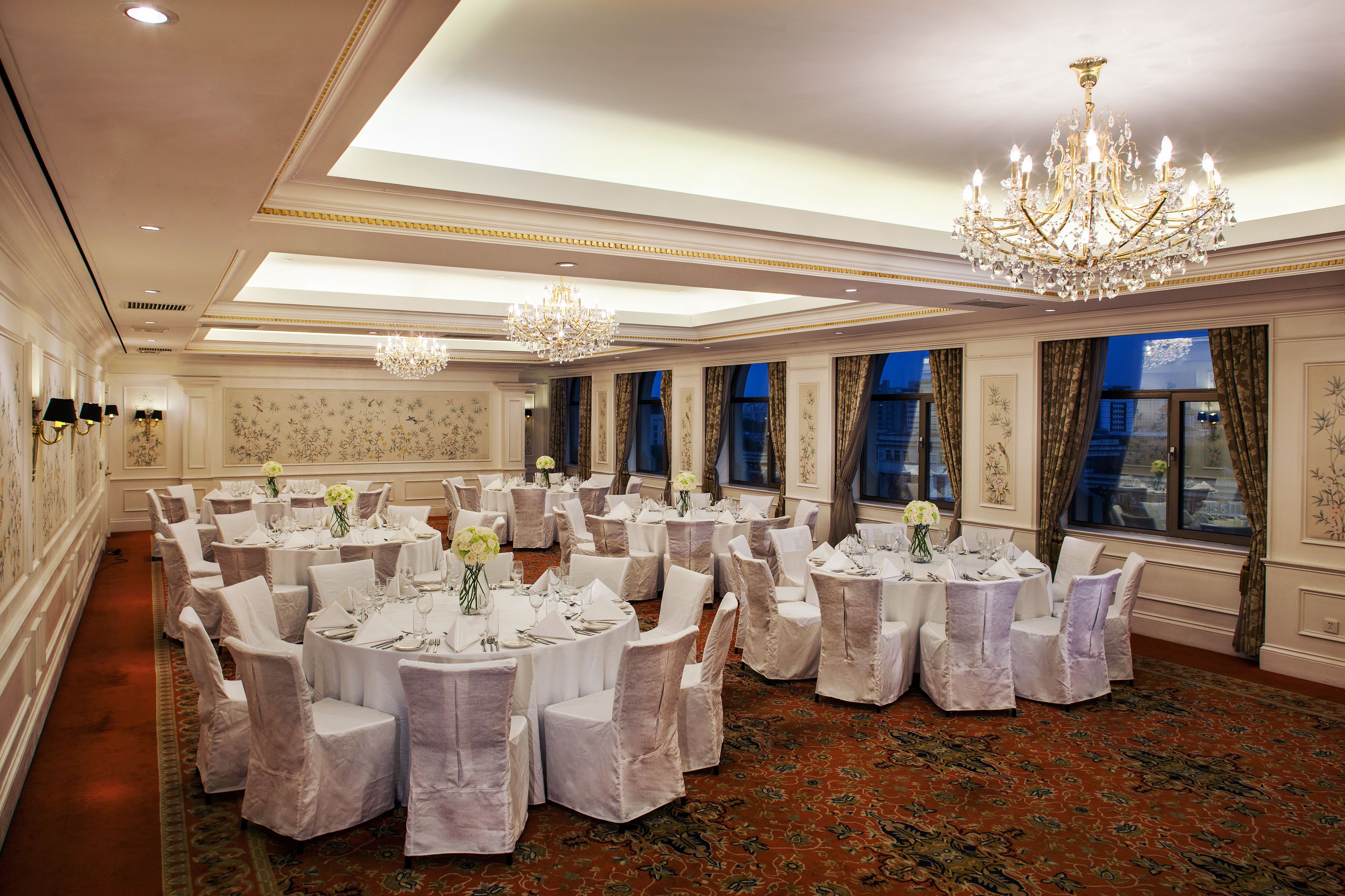 Un salón de banquetes con mesas redondas, sillas blancas, candelabros y amplias ventanas con cortinas.