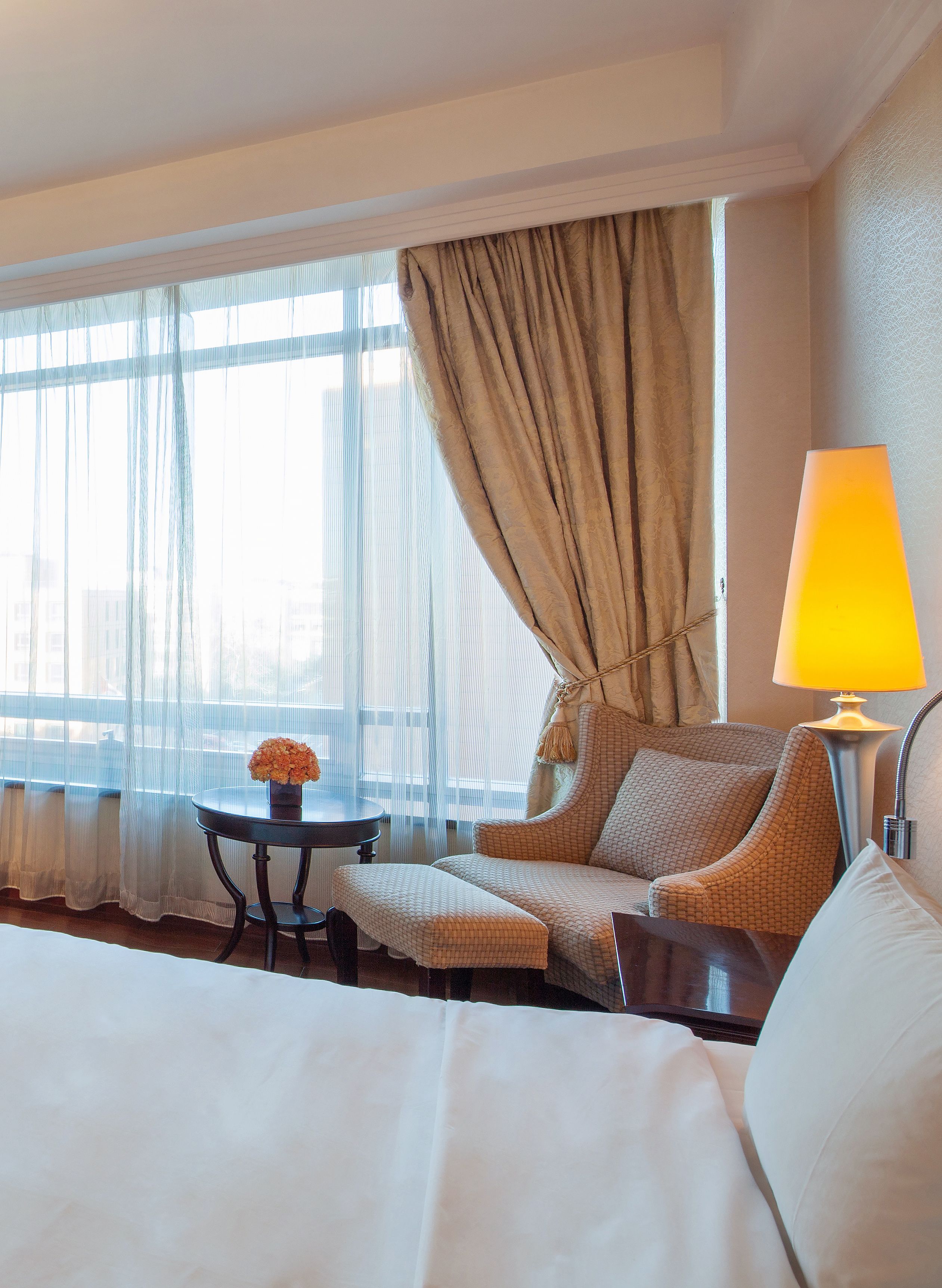 Een hotelkamer met een bed, een sofa, een kleine tafel met een bloevas, een lamp en een raam met gordijnen.