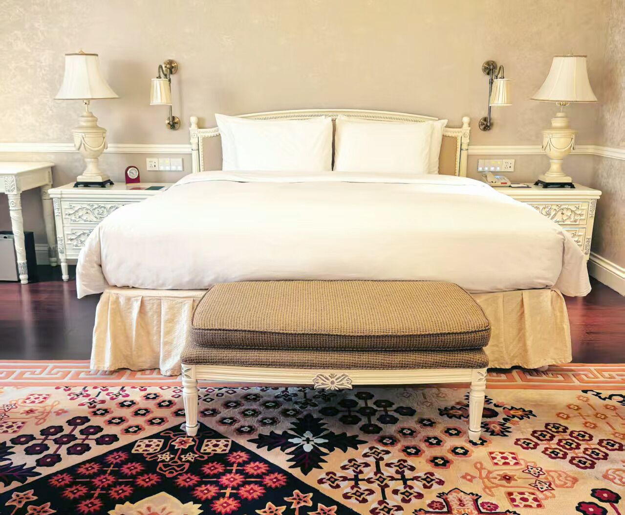 A neatly made bed with a white comforter and pillows, a wooden bench with a cushion, and two bedside tables with lamps.