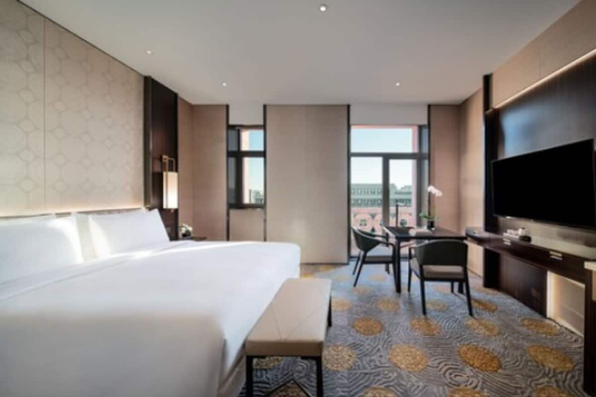 A hotel room with a large bed, a desk with a chair, a television, and a window offering a city view.