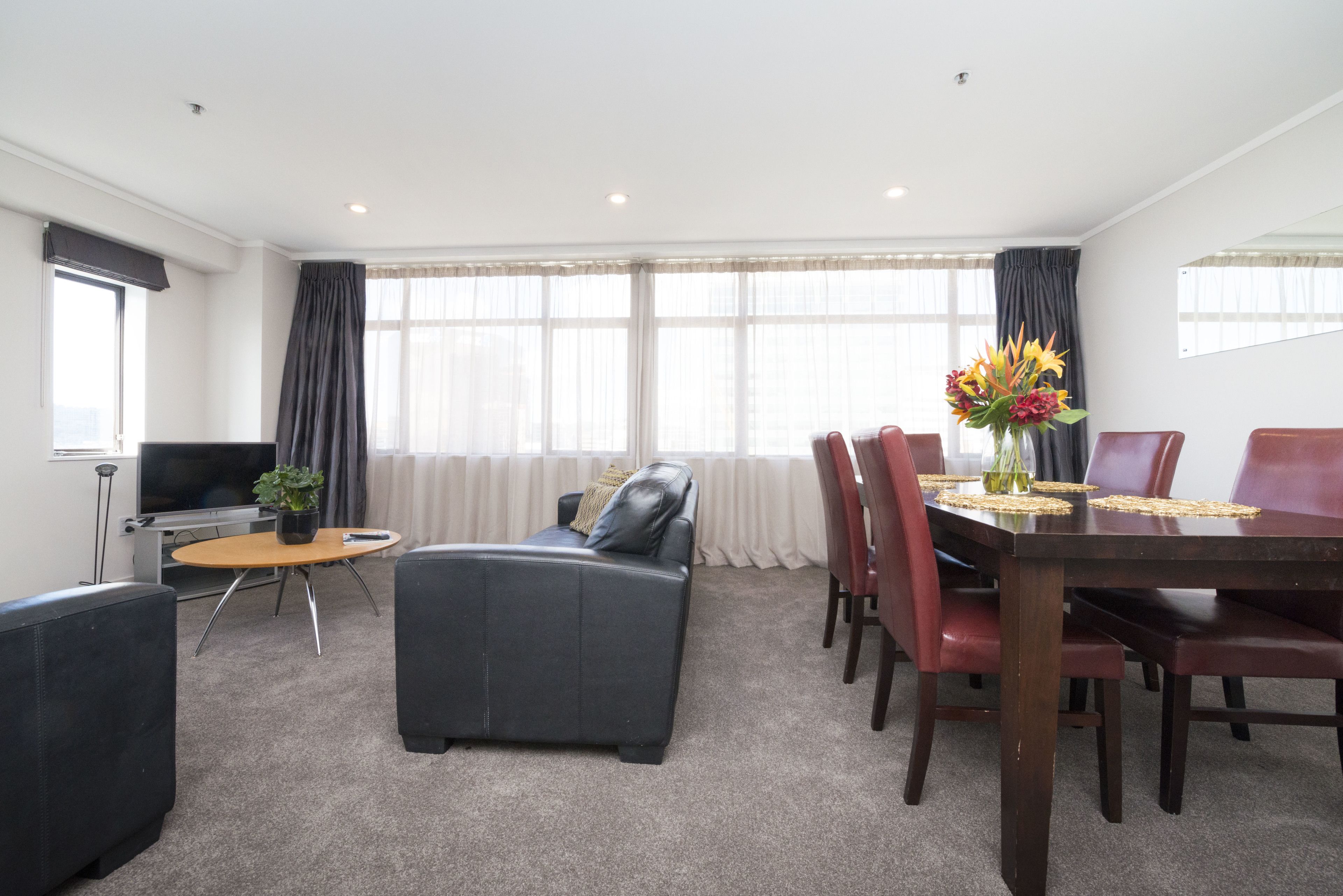 A modern hotel room with a sofa, a dining table with chairs, a TV, and a large window with curtains.