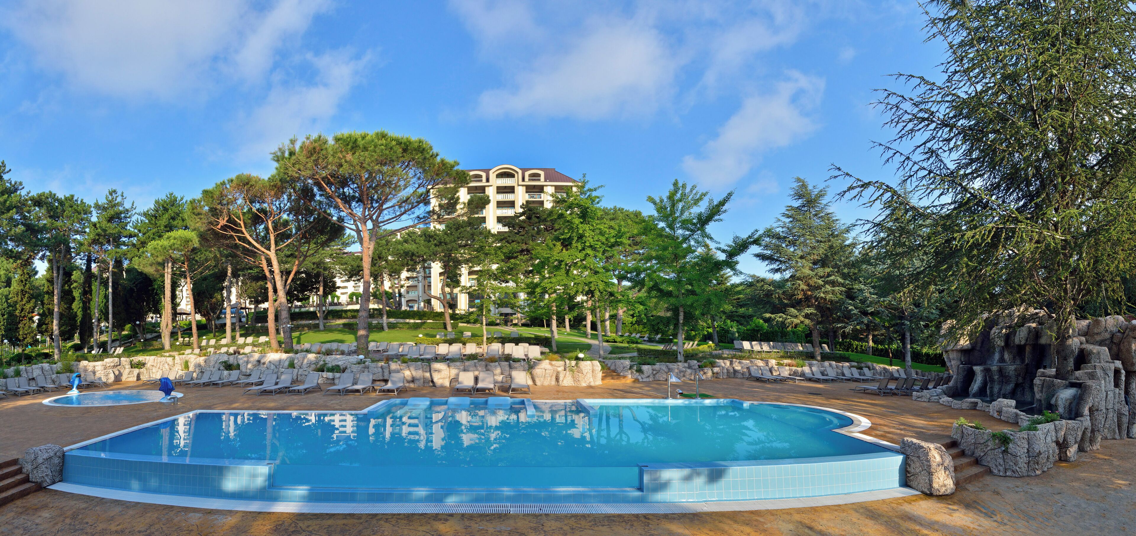 Una piscina con sillones de descanso, rodeada de árboles y un edificio al fondo.