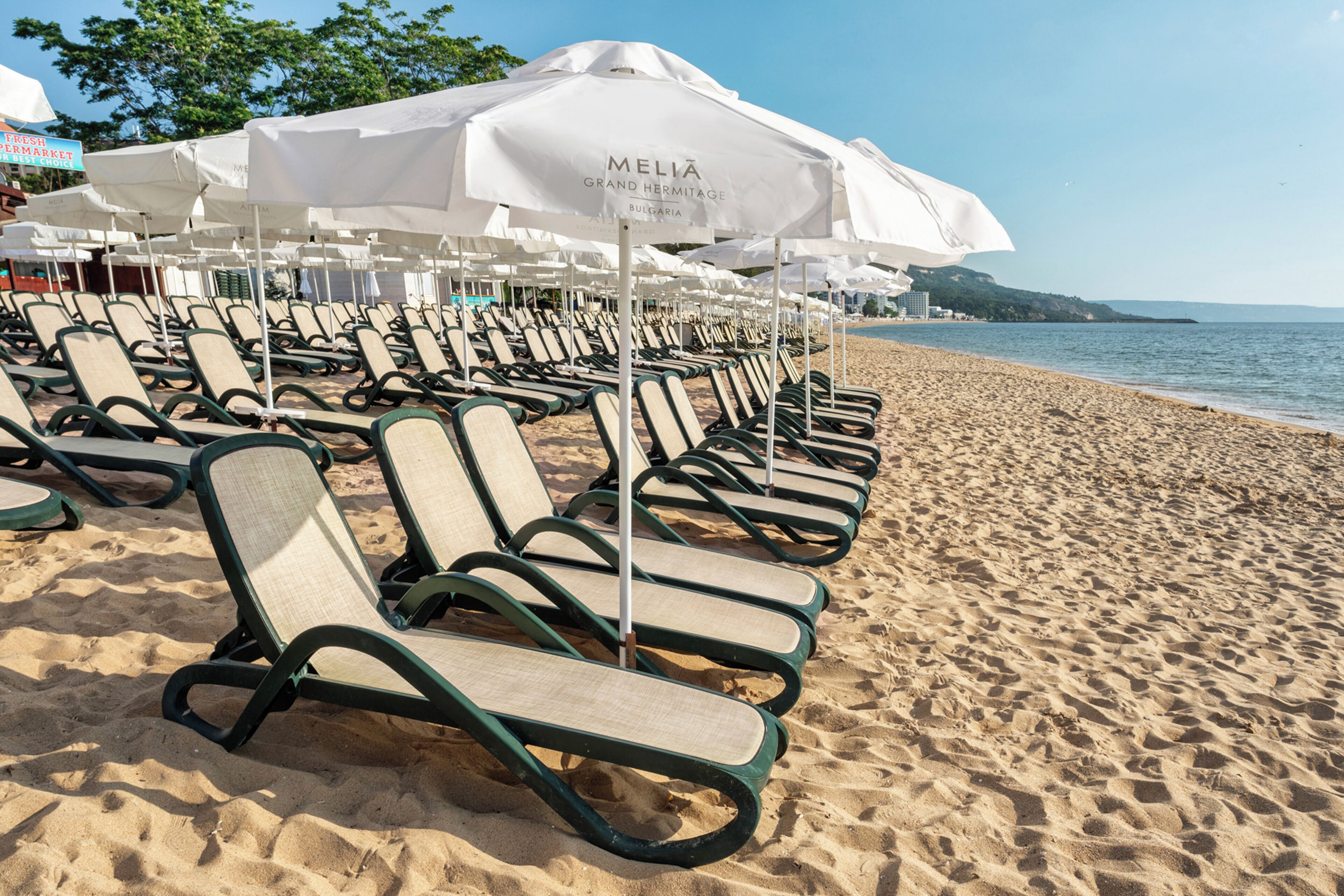 A beachside lounge area with rows of sun loungers and white umbrellas, a sandy beach, and a clear view of the sea.