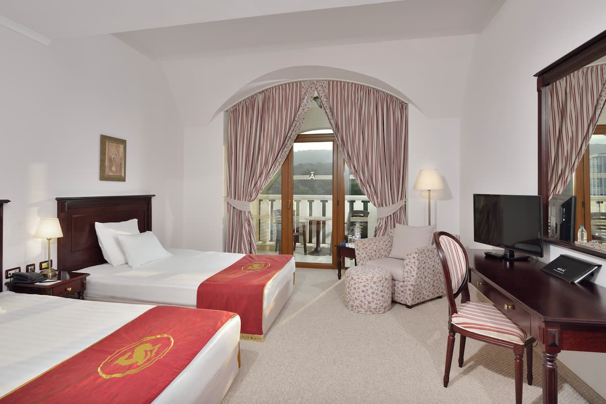 Une chambre d’hôtel avec deux lits, un bureau, une chaise, une télévision et un balcon avec des rideaux.