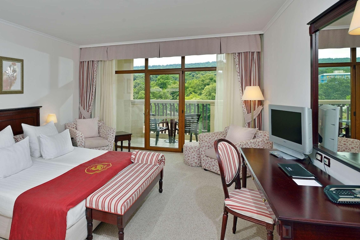 A hotel room with a bed, a desk with a computer, a TV, and a balcony with outdoor seating.