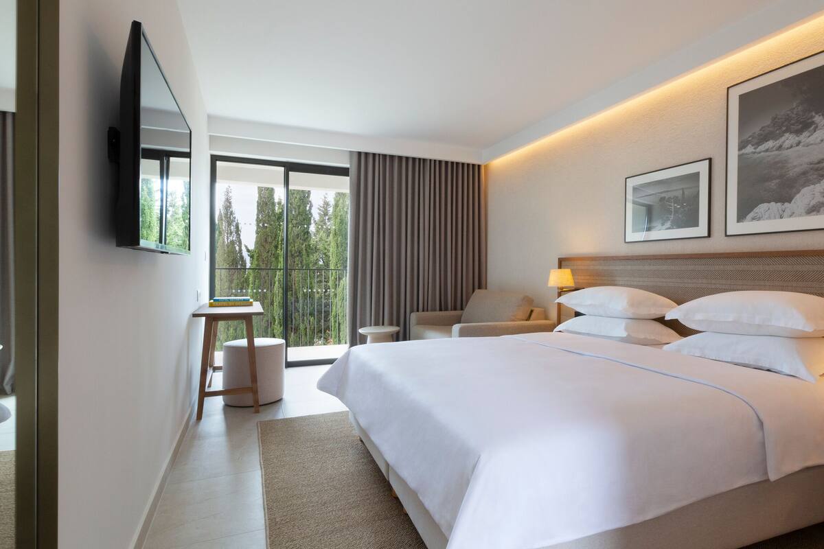 A modern hotel room with a large bed, a desk, a chair, and a view of the outdoors.
