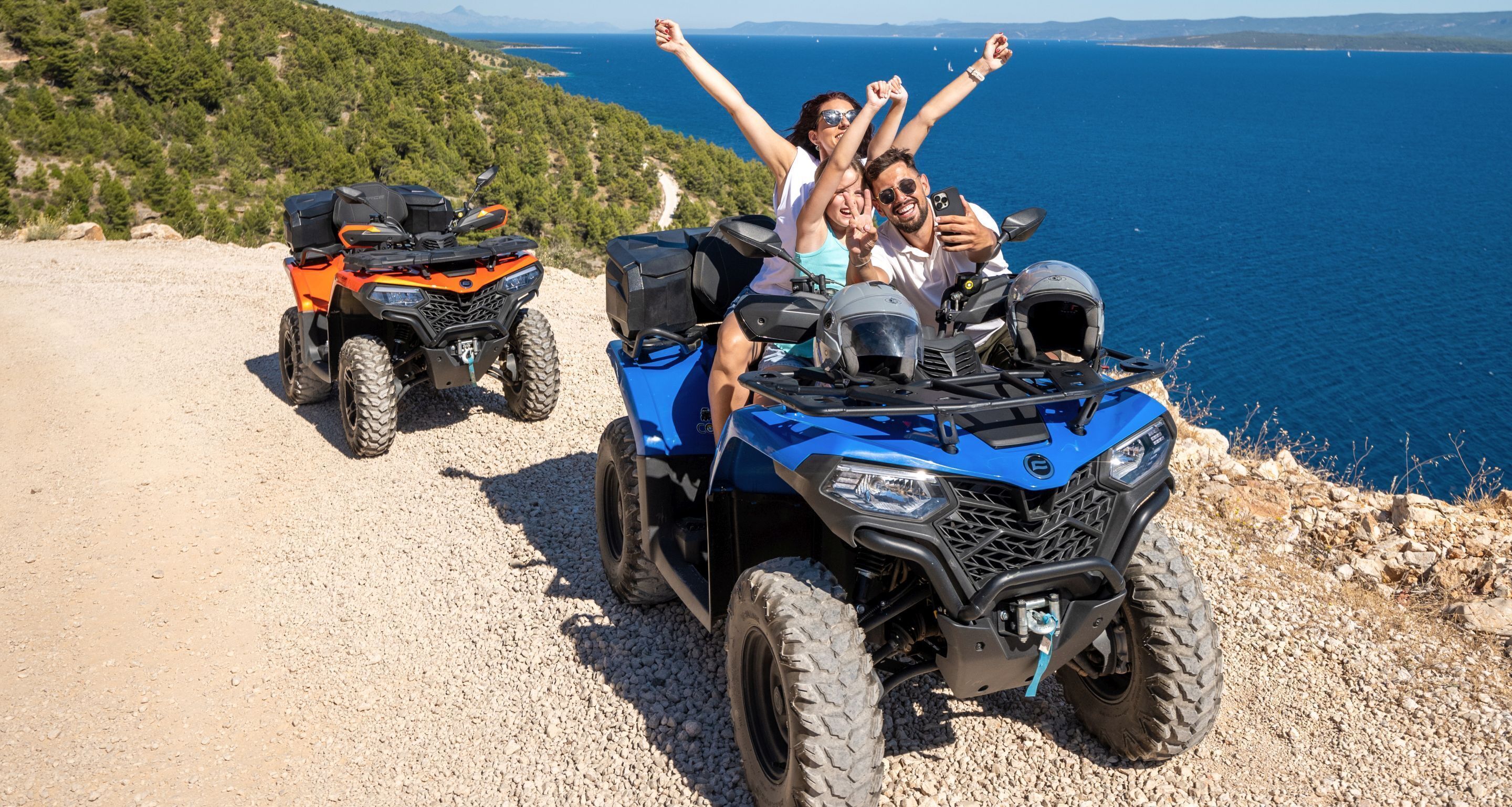 A group of people on ATVs on a gravel path with a scenic view of the sea and hills in the background.