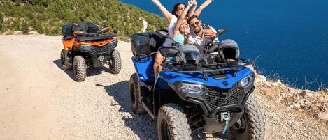A group of people on ATVs on a gravel path with a scenic view of the sea and hills in the background.