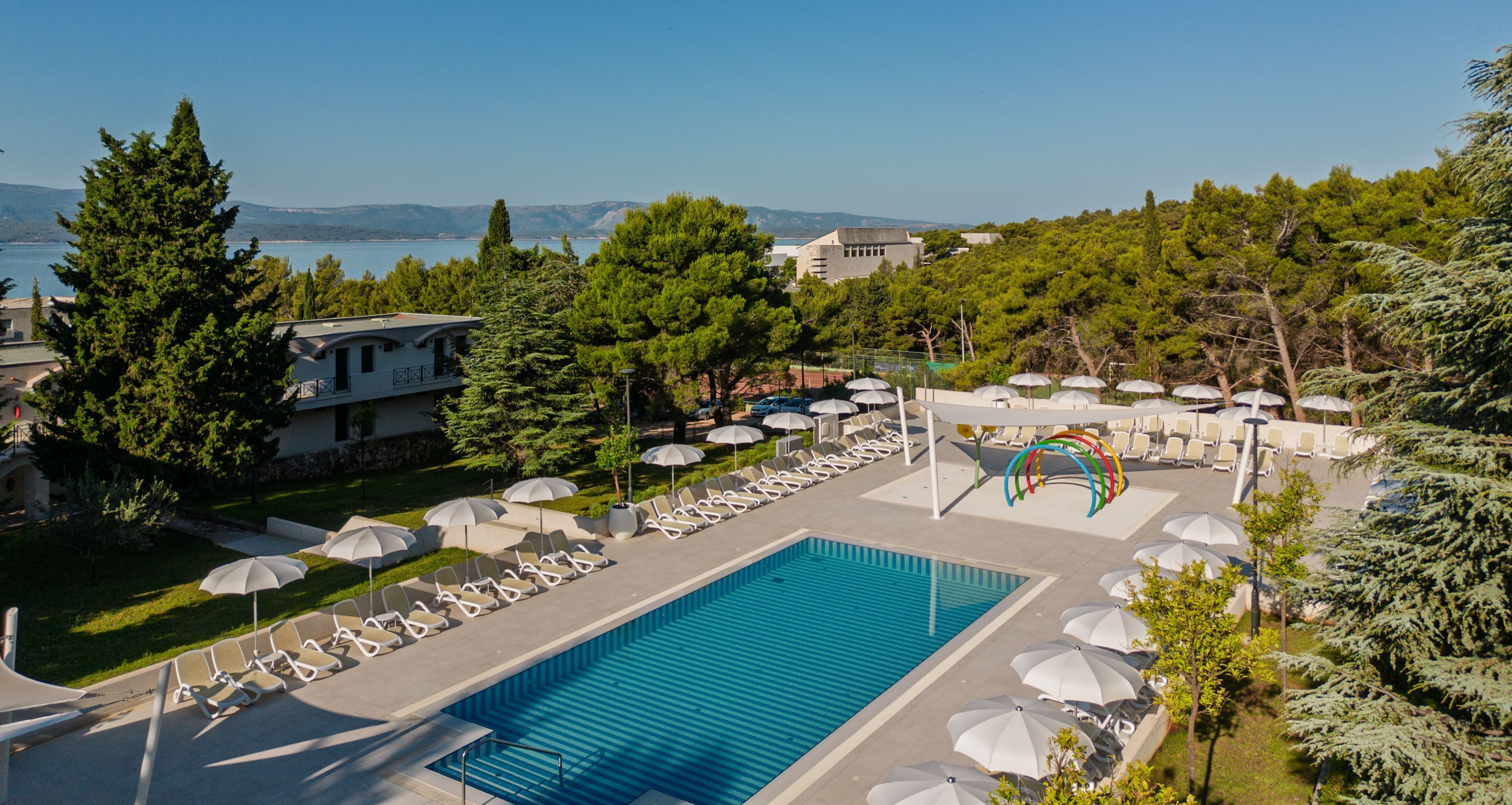 Ein Swimmingpool mit Liegestühlen und Sonnenschirmen, umgeben von Bäumen und Gebäuden.