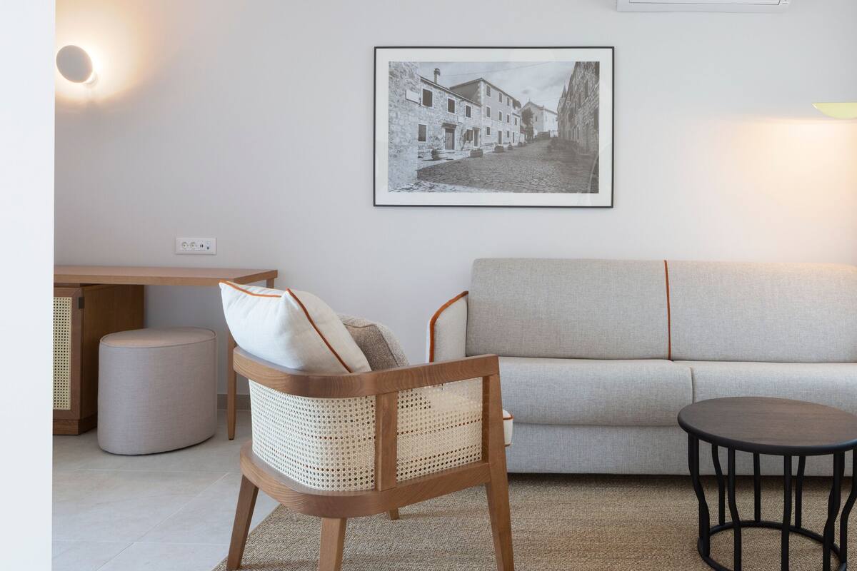 A living room with a sofa, a wooden chair, a round table, a lamp, a picture on the wall, and a small stool.