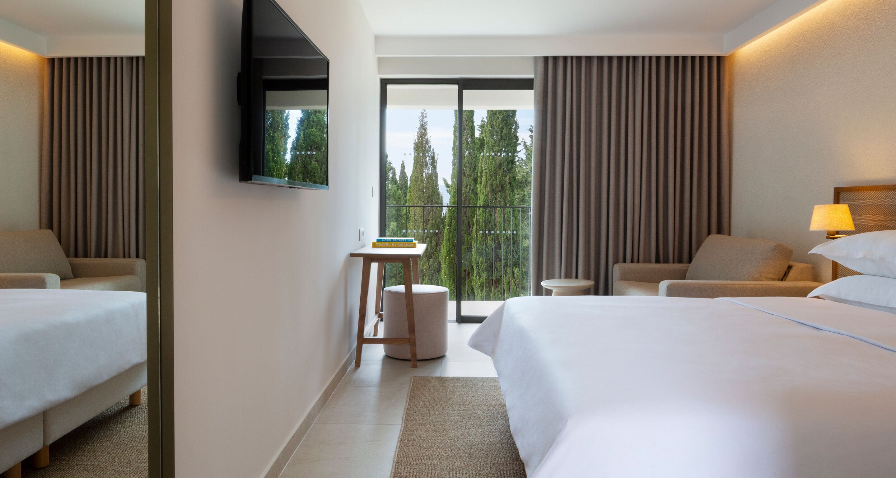 Ein modernes Hotelzimmer mit einem großen Bett, zwei Sesseln, einem kleinen Tisch und einem an der Wand befestigten Fernseher.