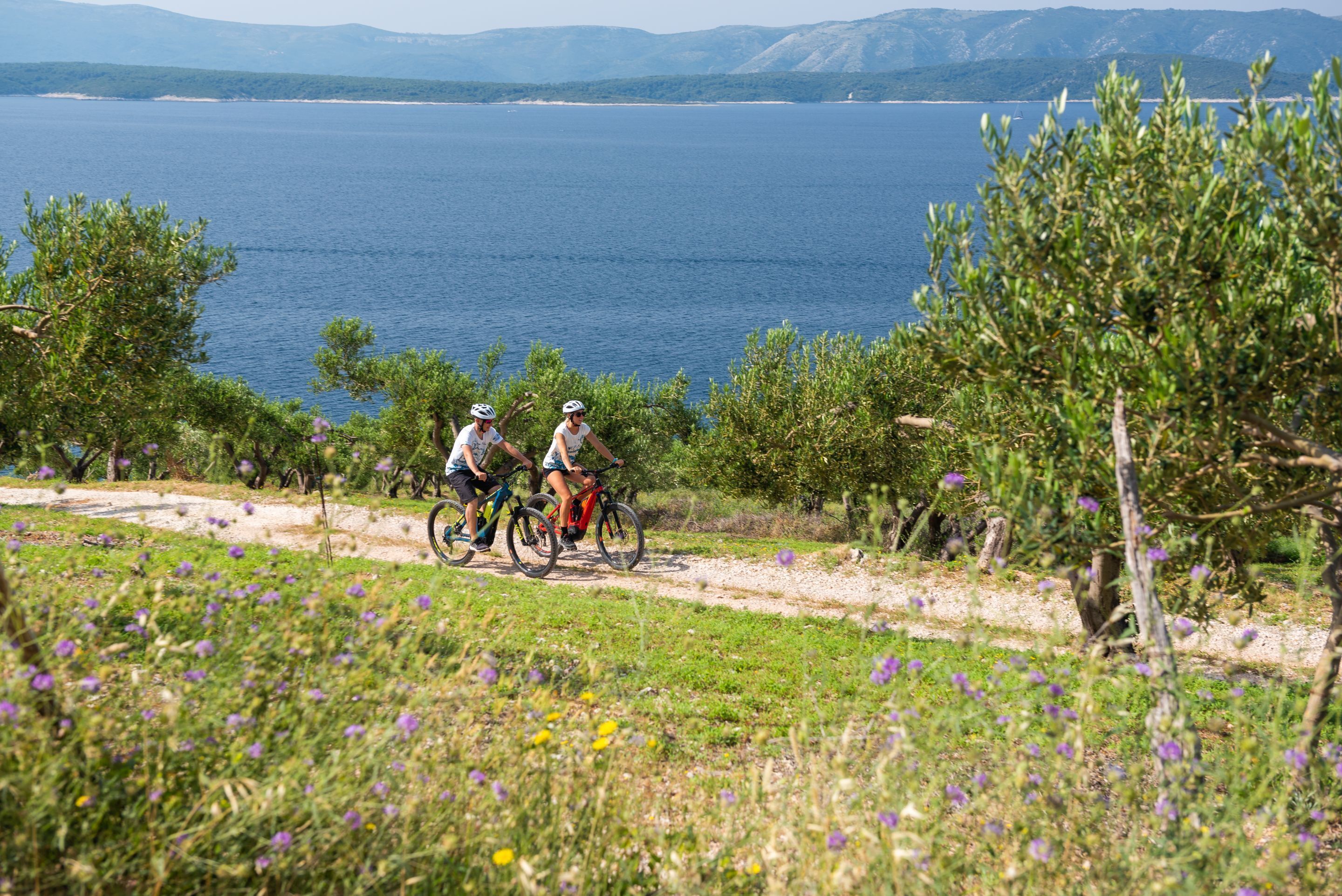 Two cyclists on a dirt path with a lake and mountains in the background.