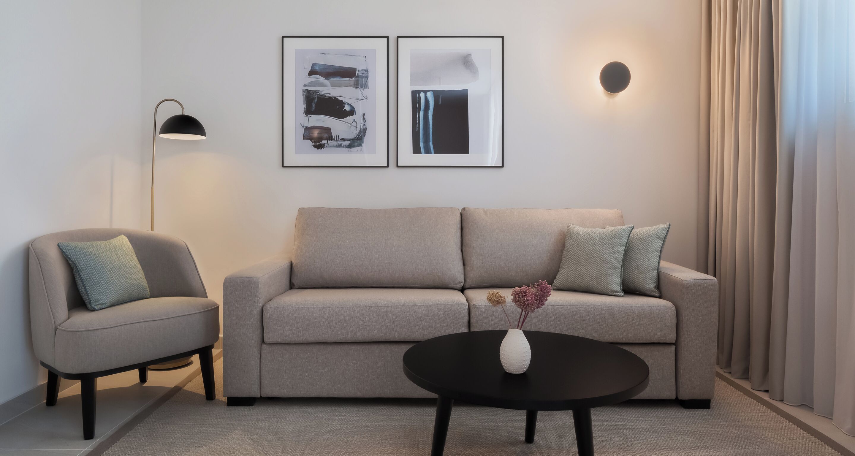 Un salon moderne avec un canapé, un fauteuil et une table basse agrémentée d’un vase. Deux œuvres d’art encadrées ornent le mur, et une lampe sur pied est placée dans un coin.