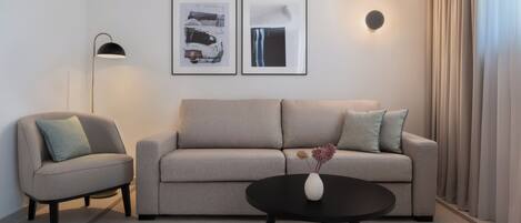 A modern living room with a sofa, armchair, and a coffee table with a vase. There are two framed artworks on the wall and a floor lamp.