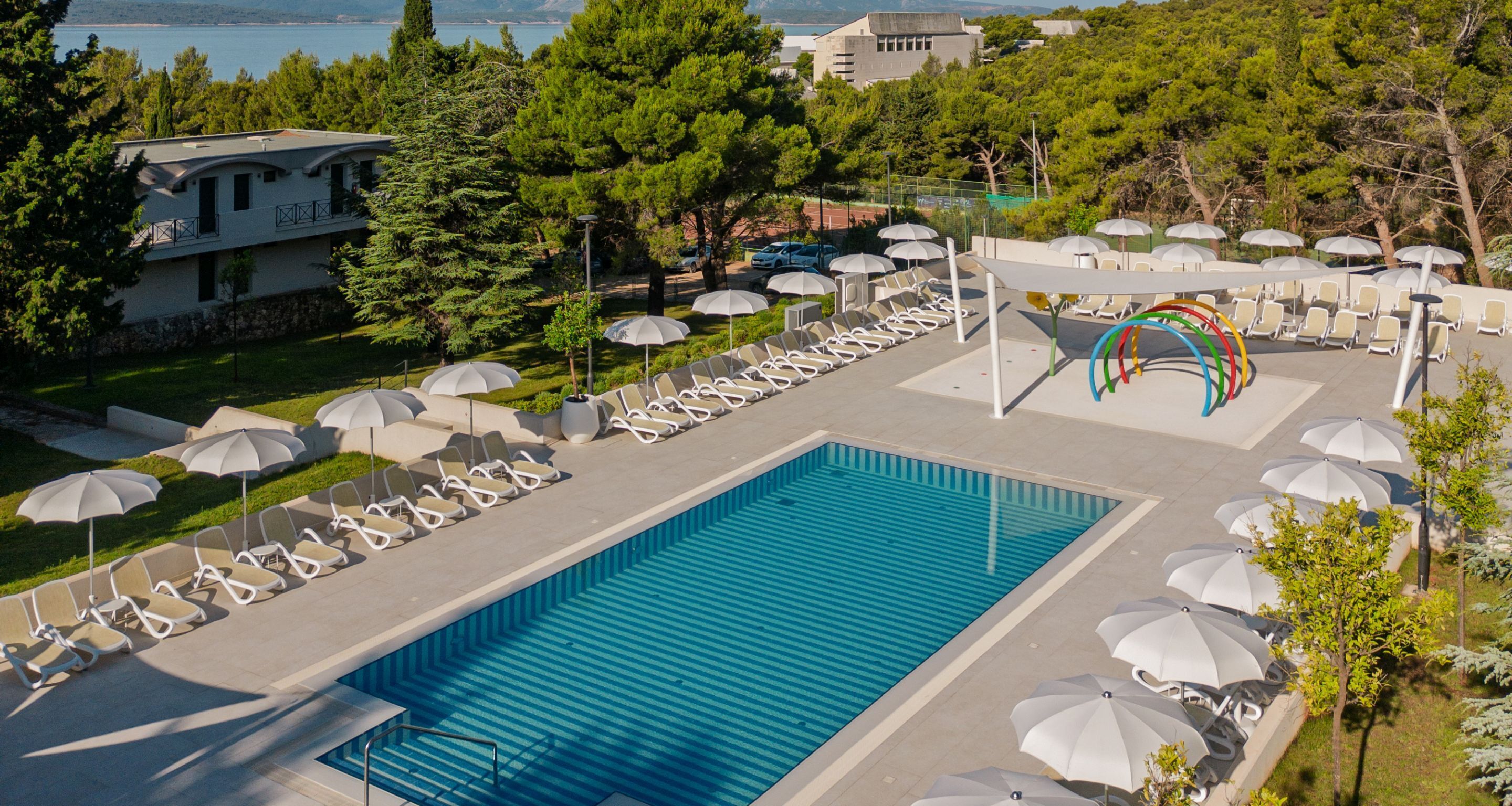 A pool area with lounge chairs and umbrellas, surrounded by trees and a view of the sea.
