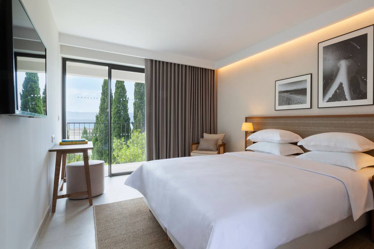 A modern hotel room with a large bed, a side table, a chair, and a view of trees through large windows.