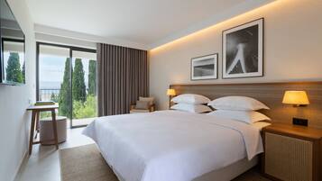 A modern hotel room with a large bed, a side table, a chair, and a view of trees through large windows.