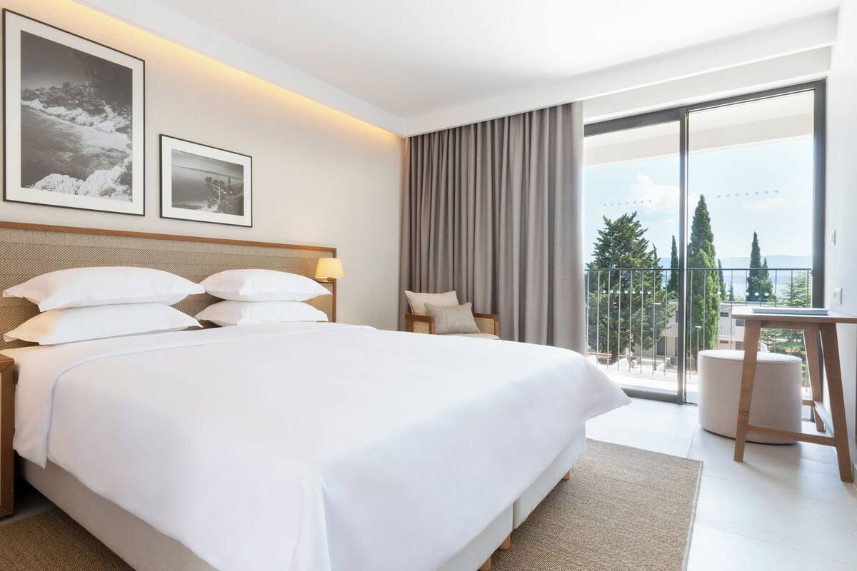 A modern hotel room with a large bed, two bedside tables with lamps, a large window with a view of trees, and a balcony.