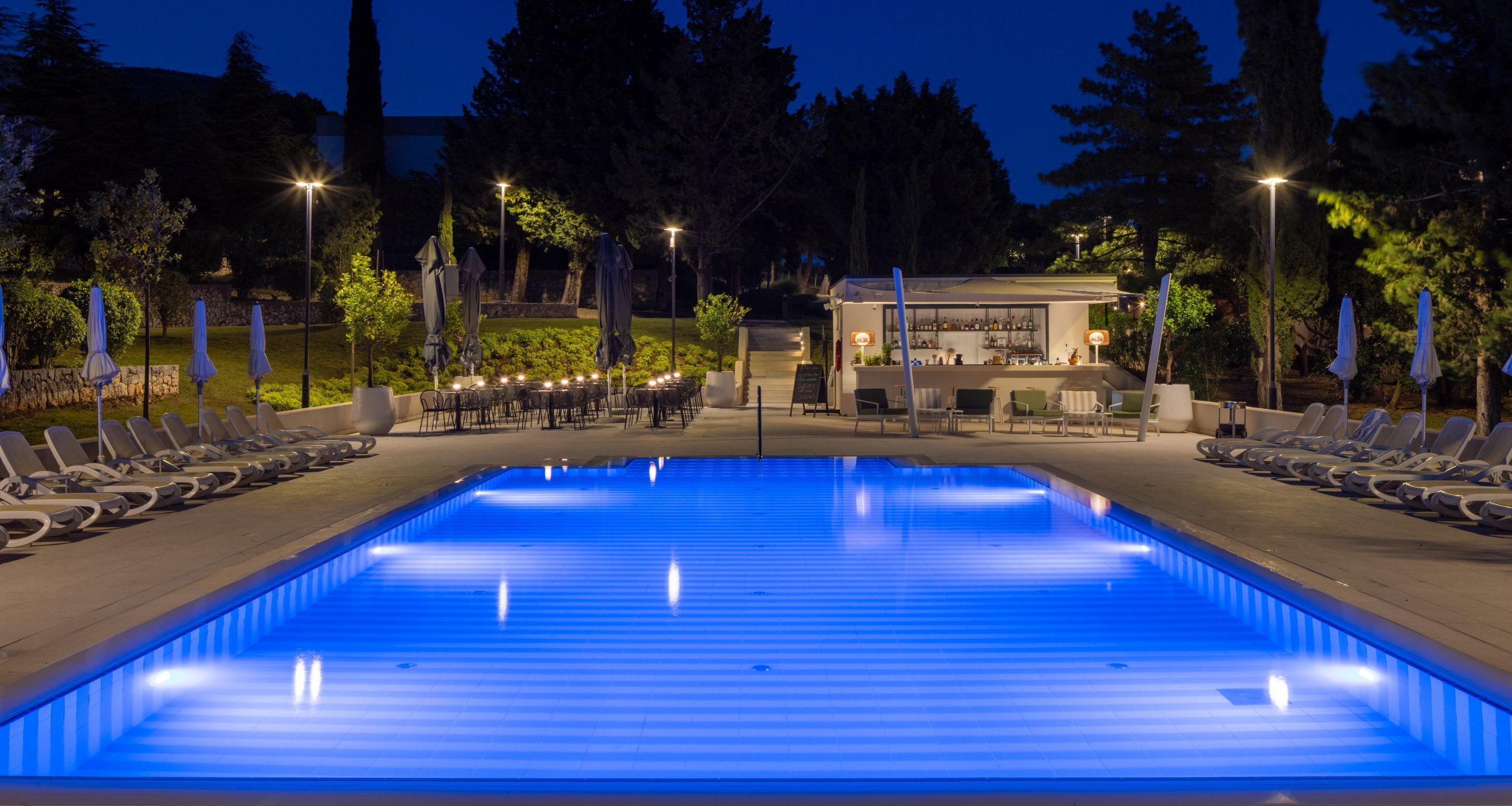 Une piscine dotée de chaises longues et d’un espace bar, le tout entouré d’arbres et éclairé la nuit.