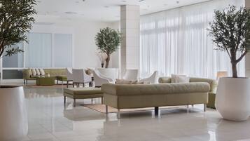 Un vestíbulo moderno con un sofá color beige, sillas blancas y plantas grandes en macetas.