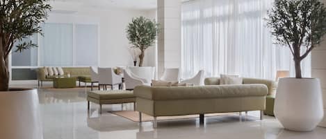 Um lobby moderno com um sofá bege, cadeiras brancas e plantas grandes em vasos.