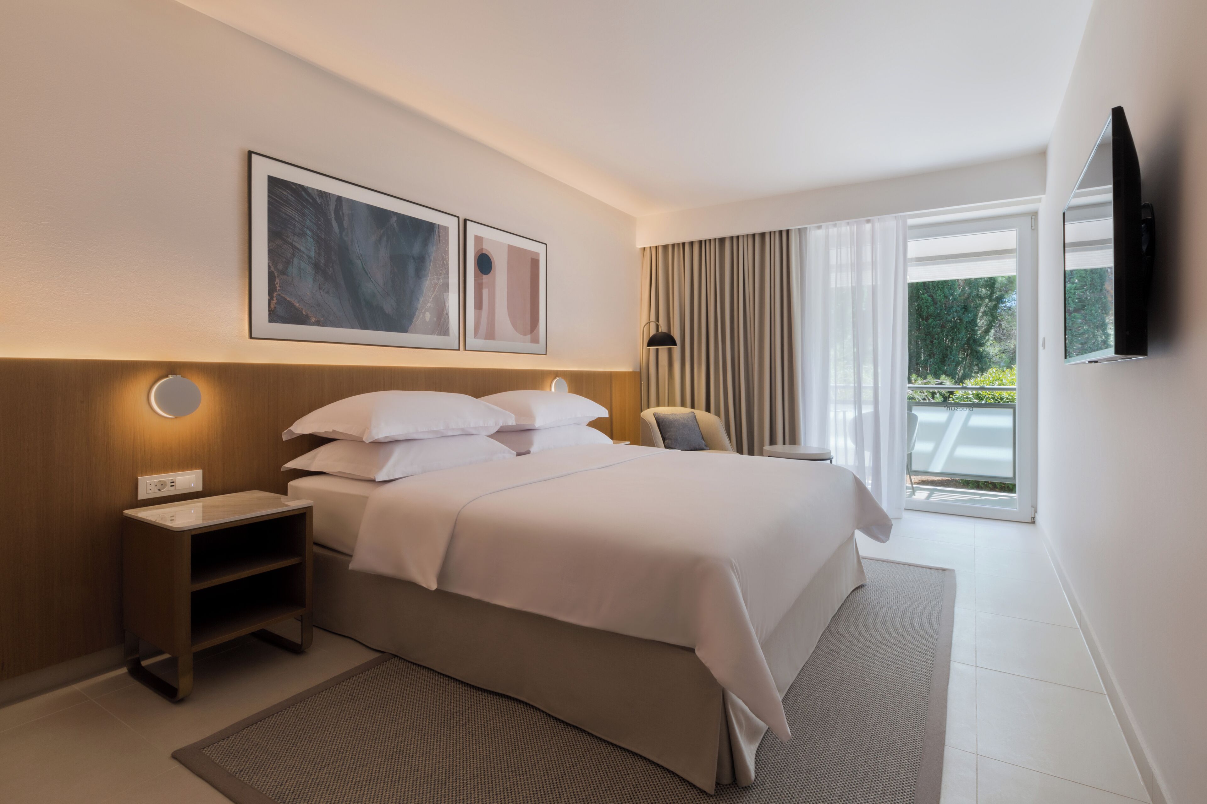 Een moderne hotelkamer met een groot bed, een nachtkastje, een flatscreen televisie aan de muur en een raam met gordijnen.