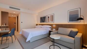 A modern hotel room with a bed, a sofa, a desk, and a chair. There are framed pictures on the wall and a lamp on the nightstand.